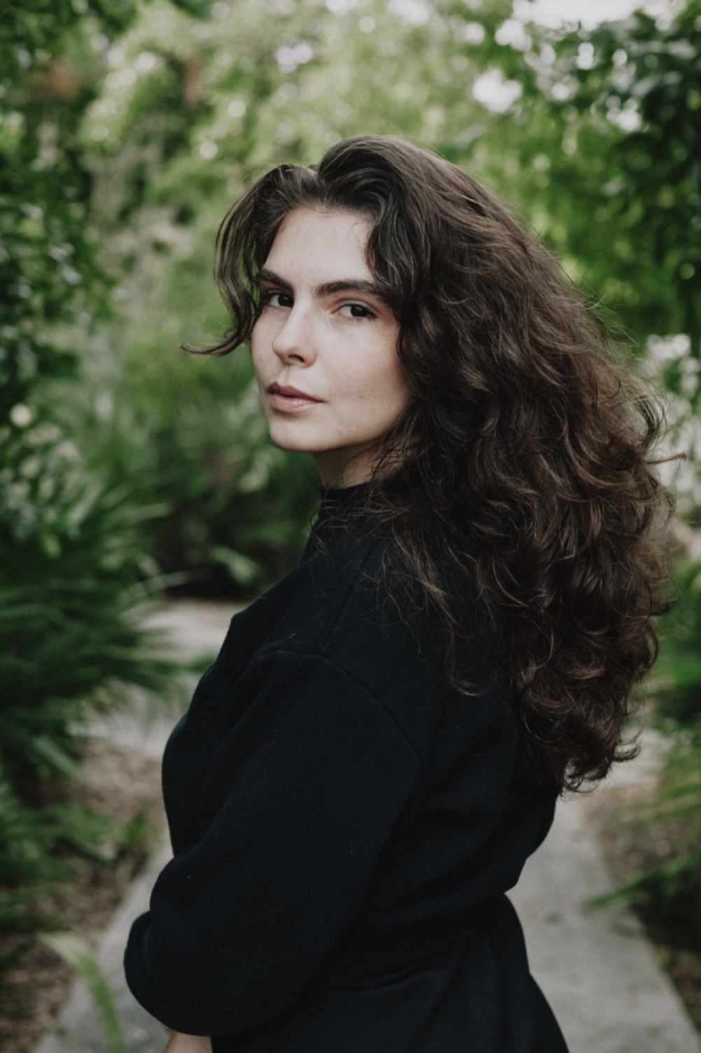 Mayra Ona, photographed outdoors among dense greenery, turning to look toward the camera